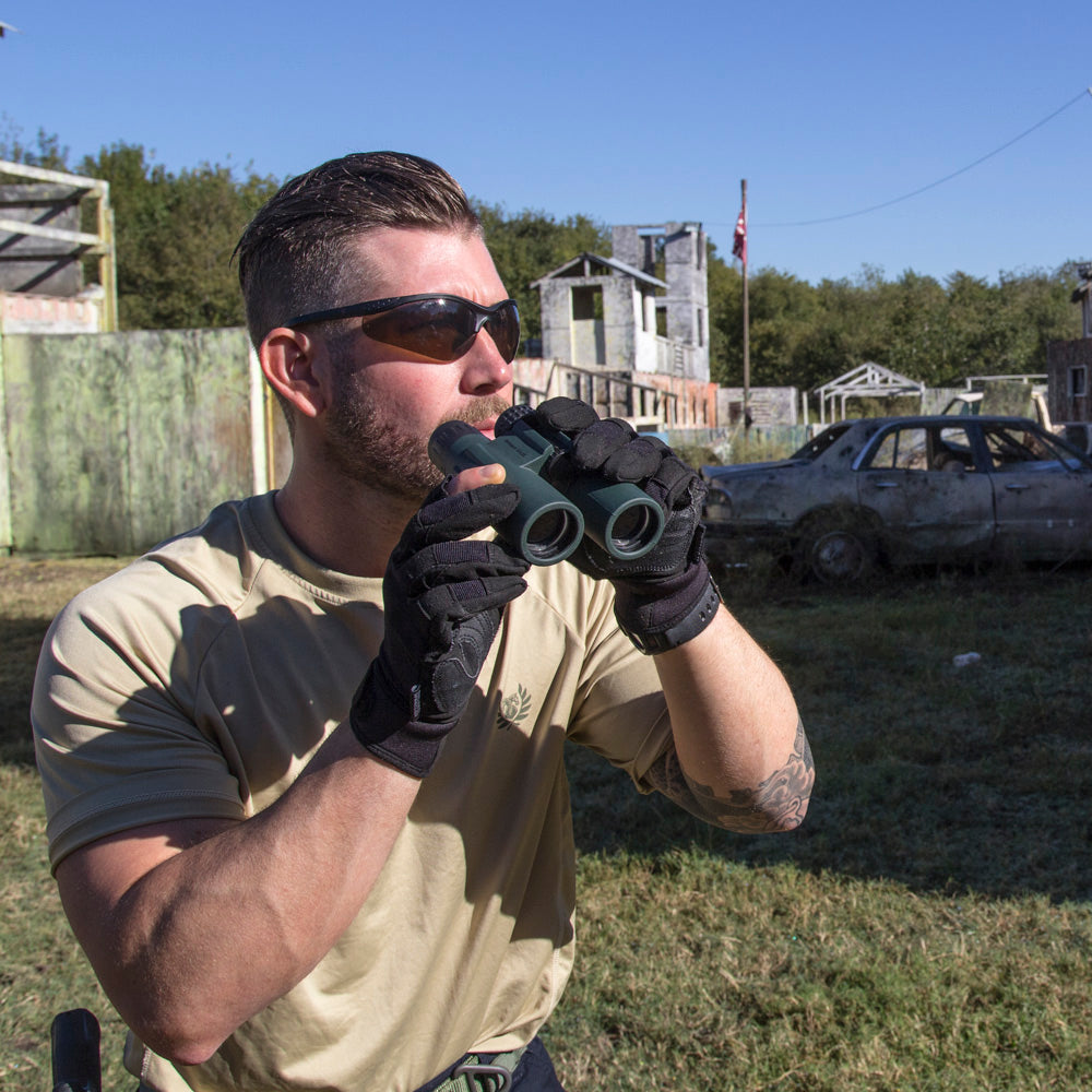 A man in tactical gear uses binoculars, observing a rundown outdoor setting with abandoned buildings and a car in the background.